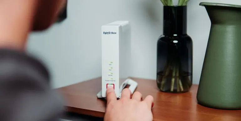 Eine Hand drückt einen roten Knopf an einer weißen FRITZ!Box auf Holz. Grüne Anzeigen leuchten. Dunkle Glas- und grüne Keramikvase im Hintergrund.