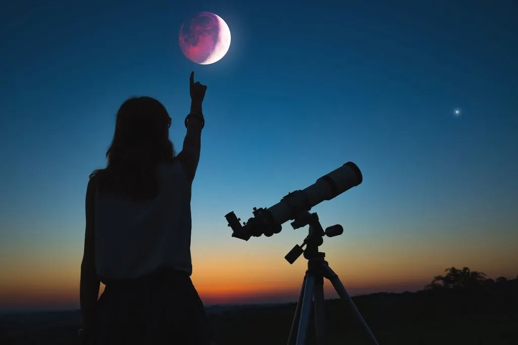 Frau zeigt auf einen pinken Mond am Abendhimmel neben einem Teleskop.
