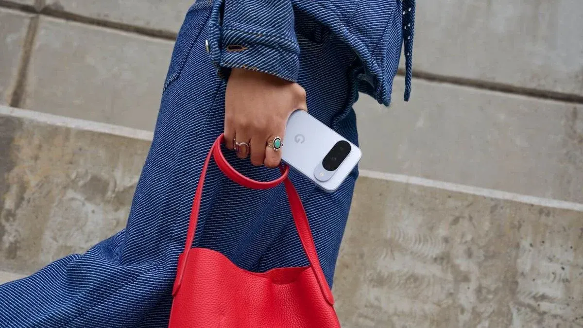 Eine Hand in blauer Jeanskleidung hält ein hellblaues Smartphone mit G-Logo, eine rote Ledertasche und trägt zwei Ringe vor Betonhintergrund.