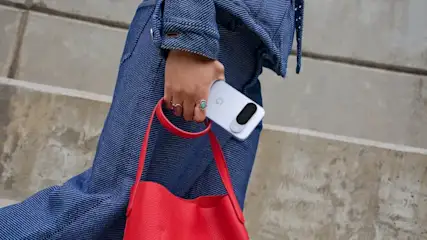 Eine Hand in blauer Jeanskleidung hält ein hellblaues Smartphone mit G-Logo, eine rote Ledertasche und trägt zwei Ringe vor Betonhintergrund.