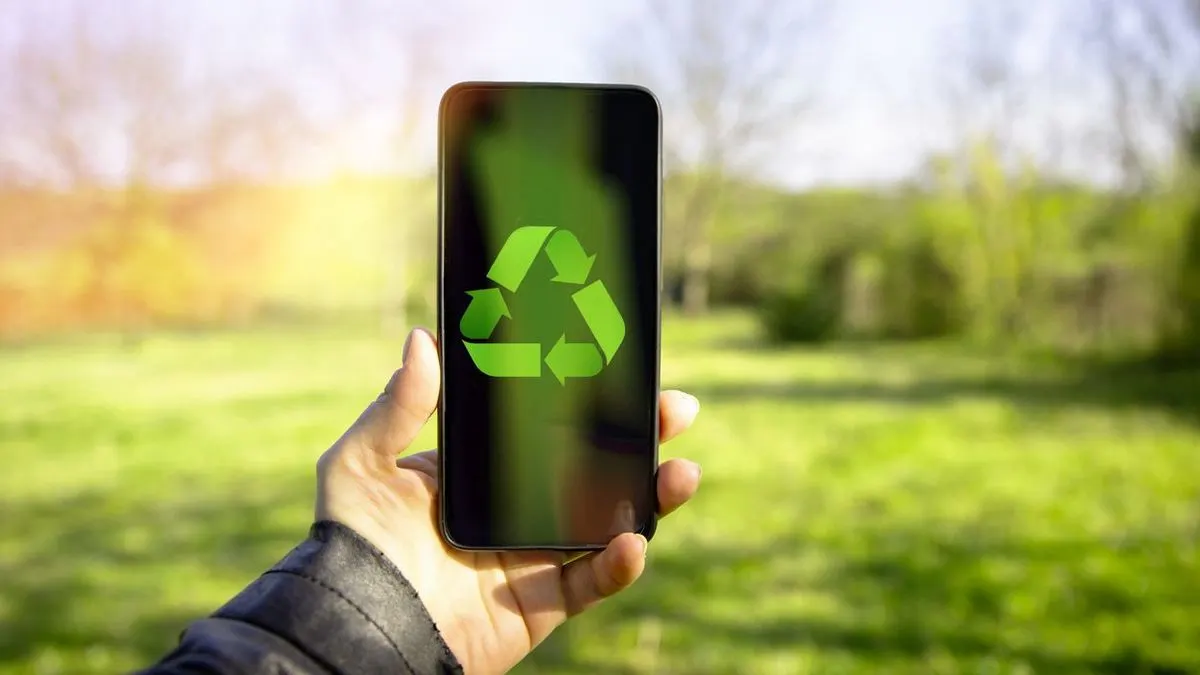 Eine Hand hält ein Smartphone mit grünem Recycling-Symbol vor unscharfem grünem Gras und Bäumen mit Sonnenlicht.