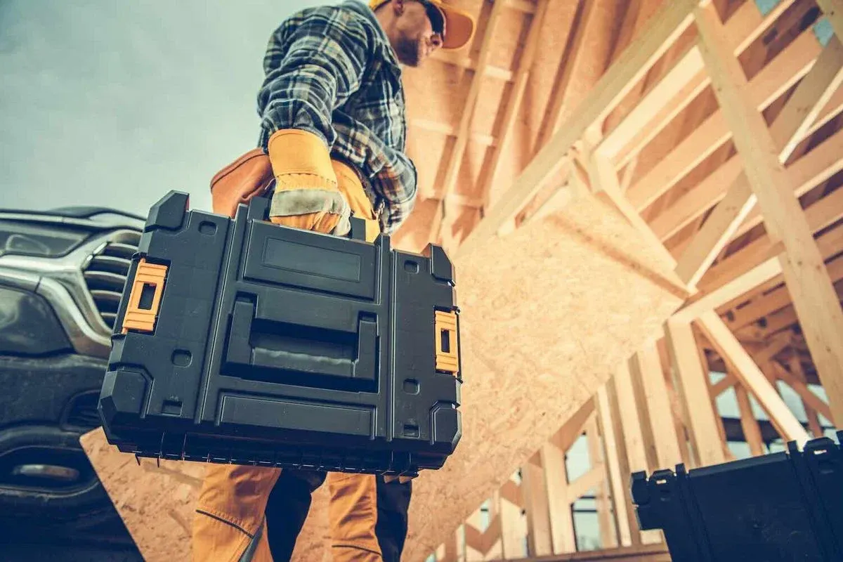 Handwerker in kariertem Hemd trägt schwarzen Werkzeugkoffer auf einer Baustelle mit Holzgerüst