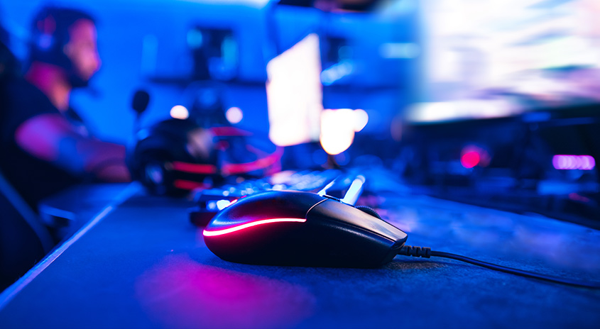 Ein schwarze Computermaus mit rotem Licht auf einem blauen Tisch. Im Hintergrund sind Bildschirme und Kopfhörer zu sehen.