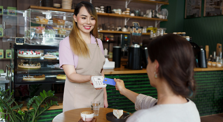  Barista reicht Kundin an einem Café-Tisch ein Kartenlesegerät, Kundin zahlt mit blauer Karte; Kaffee und Kuchen stehen auf dem Tisch.