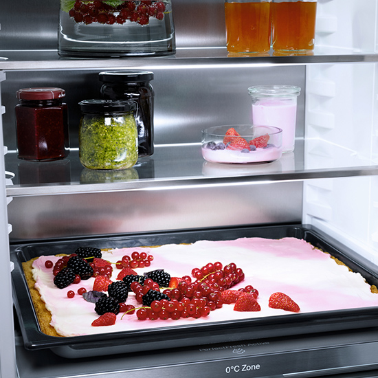 Ein Kühlschrank mit Regalen. Auf den Regalen sind Gläser mit Marmelade, Getränke und ein Backblech mit Beerenkuchen.