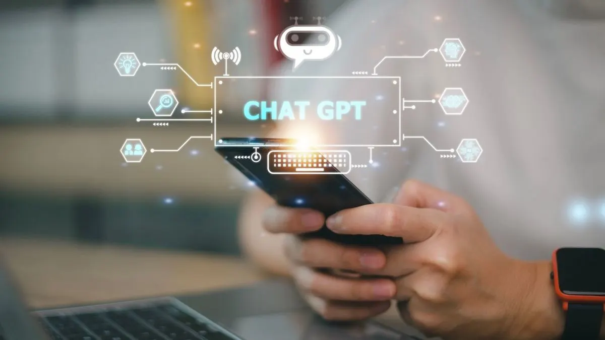 Person hält schwarzes Smartphone, darüber leuchtet "CHAT GPT" mit verbundenen KI-Symbolen. Laptop und Smartwatch sichtbar.