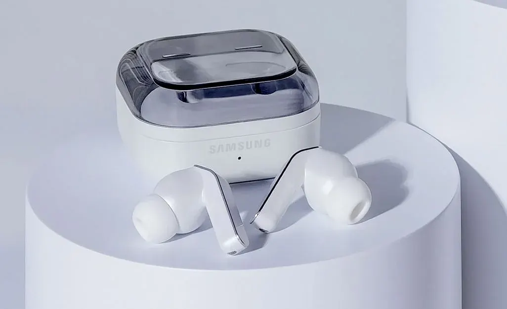Ein Paar weiße Samsung In-Ear-Kopfhörer, die vor ihrem geschlossenen Ladecase mit transparentem Deckel auf einem weißen Podest liegen.