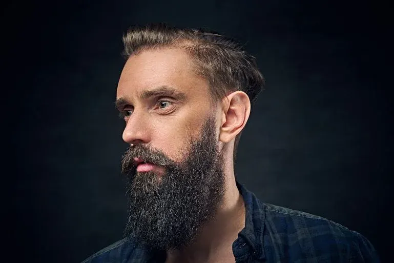 Ein Mann im Profil mit Bart und gestyltem Haar, trägt ein blaues kariertes Hemd und blickt nach links.