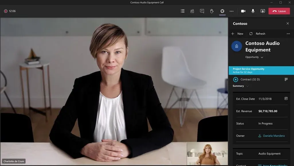 Eine Person in schwarzem Blazer und weißem Oberteil sitzt an hellem Tisch. Teams-Videoanruf mit "Contoso Audio Equipment Call" und Projektdetails (Umsatz $8,710,785.00) auf der rechten Seite. Hintergrund: weißer Stuhl, Beistelltisch.