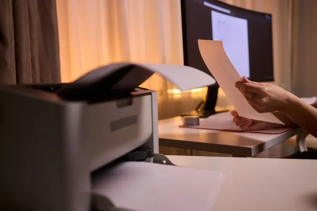 Drucker gibt Seiten aus; Person prüft gedrucktes Blatt am Schreibtisch, Monitor im Hintergrund.