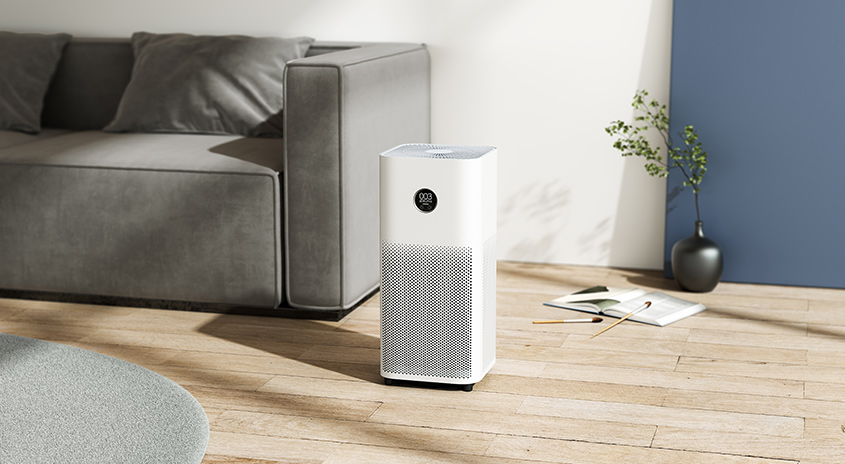 Ein weißer Luftreiniger steht auf einem Holzboden, neben einem grauen Sofa und einer Vase mit Pflanzen.