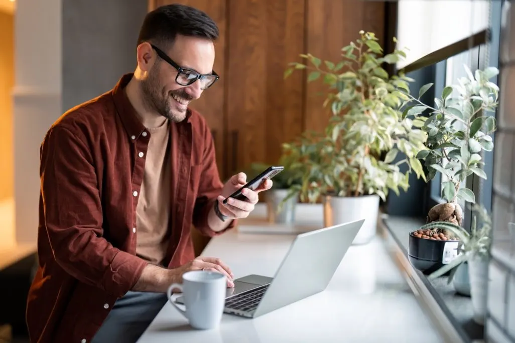 Mann sitzt an einem Schreibtisch mit Laptop und Smartphone und trinkt Kaffee.