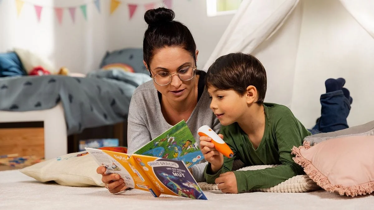 Erwachsener und Kind lesen auf einem Teppich ein illustriertes Buch. Kind hält orange-weißen Stift. Im Hintergrund: Bett, bunte Wimpelkette.