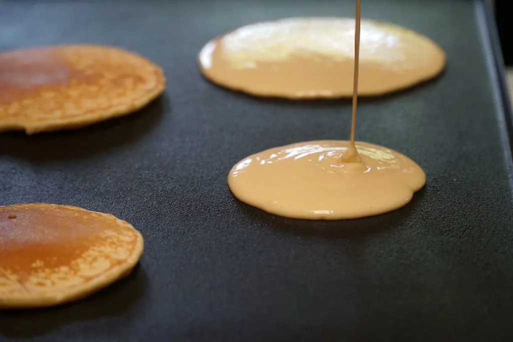 Pfannkuchenteig wird auf eine heiße Platte gegossen, daneben bereits gebackenen Pancakes.