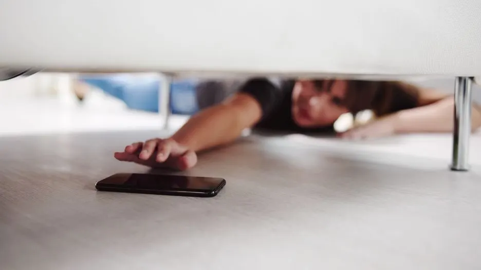 Person versucht ein Smartphone unter einem Sofa hervorzuziehen.