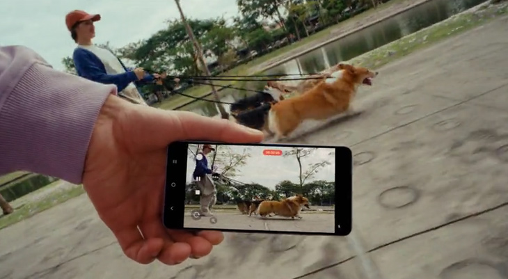 Eine Person hält ein Smartphone in der Hand und filmt jemanden, der mehrere Hunde an der Leine spazieren führt. Das Display des Smartphones zeigt die laufende Videoaufnahme mit der Szene im Hintergrund. Die Hunde laufen lebhaft und dynamisch am Ufer eines Gewässers entlang. Das Bild vermittelt den Eindruck, dass das Smartphone auch bei Bewegung stabile Videoaufnahmen ermöglicht.