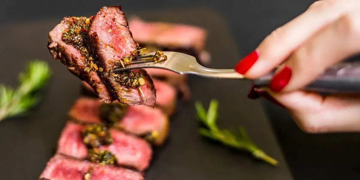 Eine Hand mit roten Nägeln hebt mit einer Gabel ein Stück medium-rare Steak mit grüner Sauce. Geschnittenes Steak und Rosmarin auf dunklem Teller.