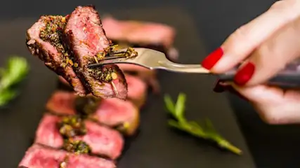 Eine Hand mit roten Nägeln hebt mit einer Gabel ein Stück medium-rare Steak mit grüner Sauce. Geschnittenes Steak und Rosmarin auf dunklem Teller.