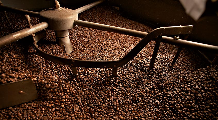 Ein großer Haufen gerösteter Kaffeebohnen wird von einer Metallvorrichtung mit Griffen und Rührarmen durchmischt.