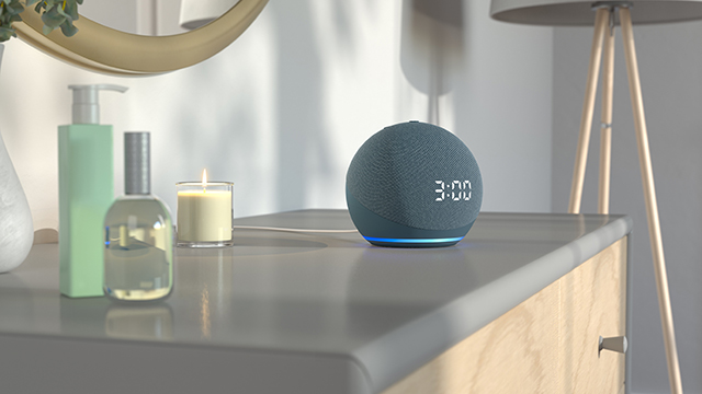 Auf einem Tisch stehen eine grüne Flasche, ein Parfum, eine Kerze und ein grauer, runder digitaler Lautsprecher.