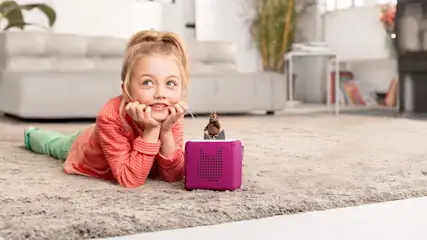 Mädchen liegt auf einem Teppich und hört lächelnd einer pinken Toniebox zu.