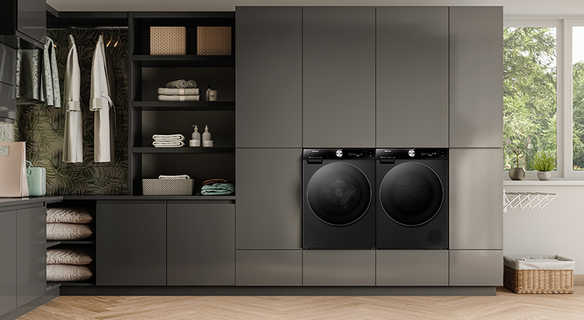 Waschküche mit zwei schwarzen Waschmaschinen, Regalen, Schränken und einem Fenster mit Blick auf Bäume.