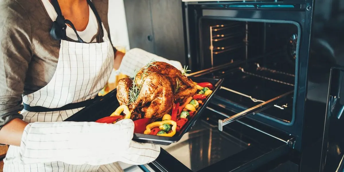 Eine Person mit karierter Kochschürze und Ofenhandschuhen nimmt ein Backblech mit gebratenem Truthahn und buntem Gemüse aus dem Ofen