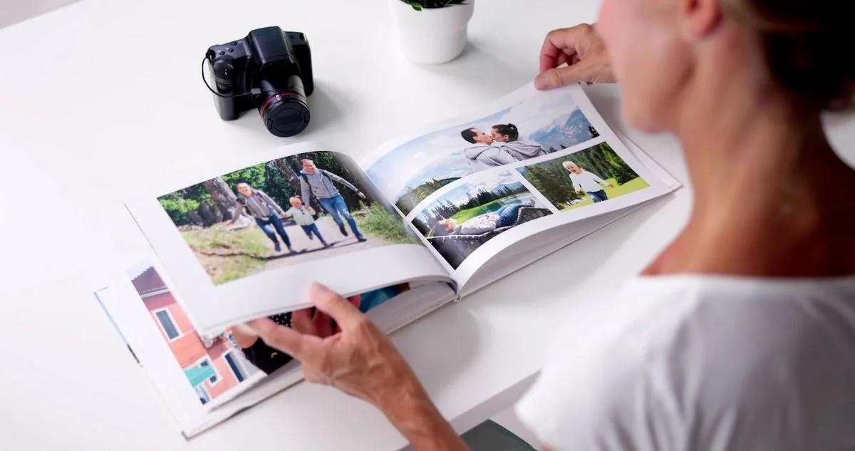 Nahaufnahme einer Person, von hinten gesehen, die auf einem weißen Tisch ein aufgeschlagenes Fotobuch mit mehreren Familienbildern (Spaziergang, Paar, Kind) durchblättert; links auf dem Tisch liegt eine Kamera, weiter hinten steht ein kleiner Blumentopf.