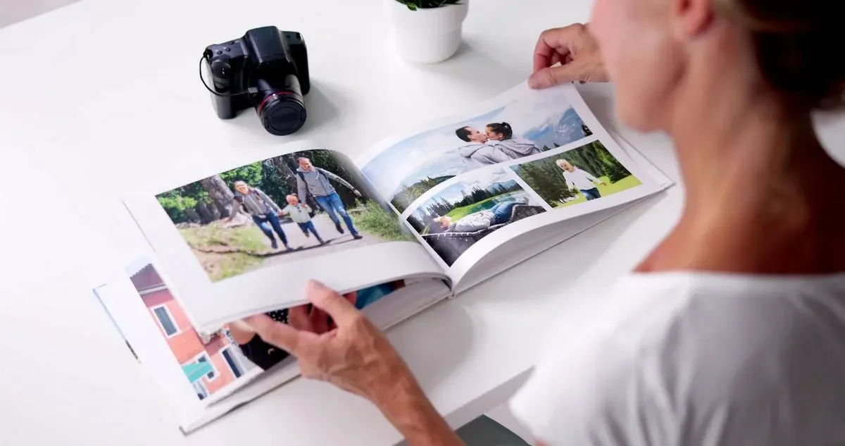 Nahaufnahme einer Person, von hinten gesehen, die auf einem weißen Tisch ein aufgeschlagenes Fotobuch mit mehreren Familienbildern (Spaziergang, Paar, Kind) durchblättert; links auf dem Tisch liegt eine Kamera, weiter hinten steht ein kleiner Blumentopf.
