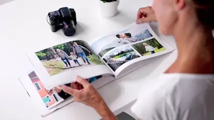 Nahaufnahme einer Person, von hinten gesehen, die auf einem weißen Tisch ein aufgeschlagenes Fotobuch mit mehreren Familienbildern (Spaziergang, Paar, Kind) durchblättert; links auf dem Tisch liegt eine Kamera, weiter hinten steht ein kleiner Blumentopf.