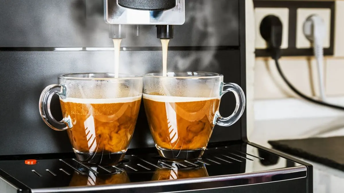 Kaffee fließt aus einer silber-schwarzen Maschine in zwei klare Gläser mit Crema. Dampf steigt auf. Im Hintergrund: Steckdose und helle Fliesenwand.