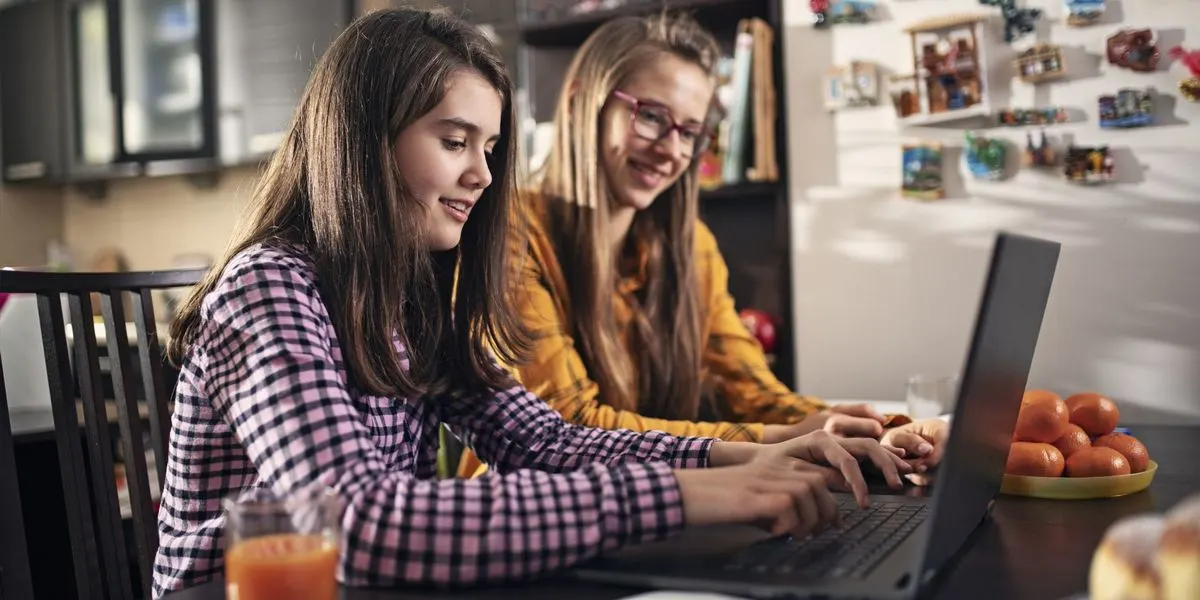 Zwei Mädchen nutzen einen Laptop zusammen am Küchentisch.