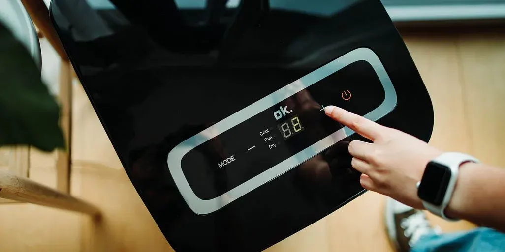Hand mit weißer Smartwatch drückt '+' Taste auf schwarzem Gerät. Bedienfeld zeigt 'ok.', '8.8', Modus und leuchtendes Einschaltsymbol. Holzboden sichtbar.