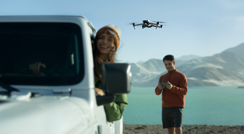 Ein weißes Auto steht vor einer Landschaft mit einem See und Bergen, während eine Drohne fliegt.