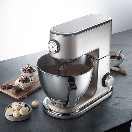 Silberne Küchenmaschine mit Schüssel steht auf einer Arbeitsfläche. Muffins, Schokolade und eine Schale mit Keksen daneben.