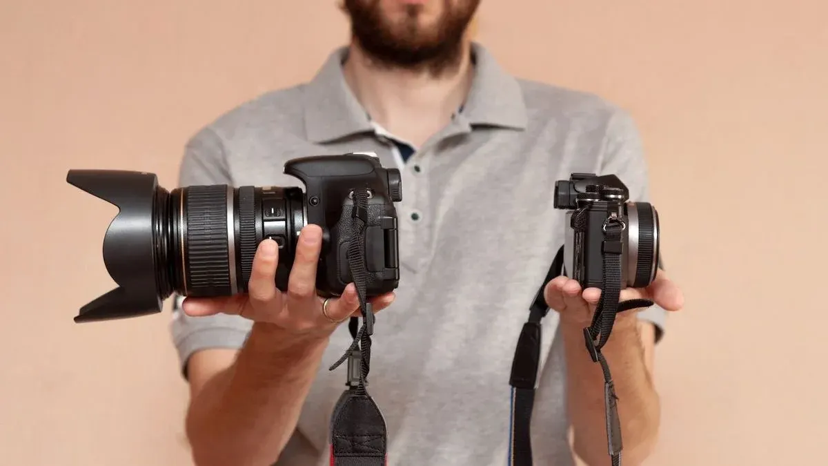 Eine Person in grauem Poloshirt hält zwei Kameras. Links eine große schwarze DSLR mit Objektiv und Gegenlichtblende, rechts eine kleinere Spiegellose.