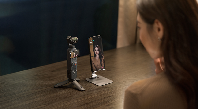 Frau sitzt vor DJI Osmo Pocket 3 Kamera-Gimbal und Smartphone auf Ständer. Smartphone zeigt Videoaufnahme der Frau. Setup für Content Creation und Streaming.