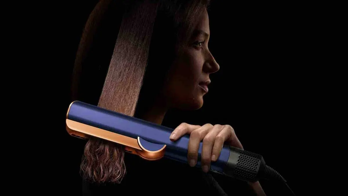 Frau glättet mit einem blauen und goldenen Dyson Airstrait Haarstyler ihr langes braunes Haar
