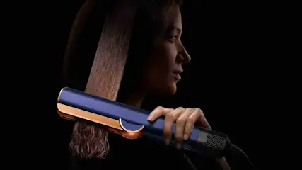 Frau glättet mit einem blauen und goldenen Dyson Airstrait Haarstyler ihr langes braunes Haar