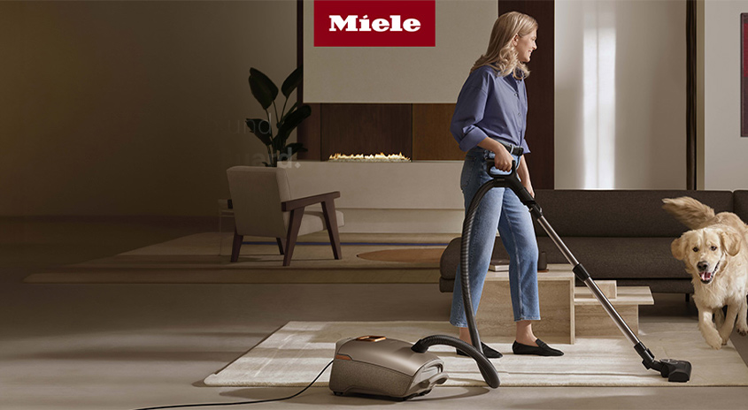 Eine Frau benutzt einen grauen Staubsauger auf einem hellen Teppich. Ein Hund steht im Hintergrund neben einem Sofa. Das Miele-Logo ist oben zu sehen.
