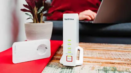 Weißer FRITZ!Box Router und MAGIC LAN Adapter auf rotem Tuch auf Holztisch. Pflanze und Person am Laptop im Hintergrund.