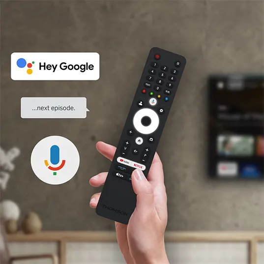 Hand hält eine schwarze TV-Fernbedienung. Auf dem Bildschirm im Hintergrund läuft ein Programm. "Hey Google"- und Mikrofon-Symbol.