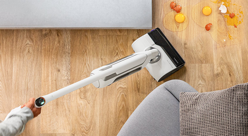 Akku-Staubsauger wischt zerbrochene Eier vom Holzboden neben Sofa und Teppich, Hand führt das Gerät für schnelle Reinigung.