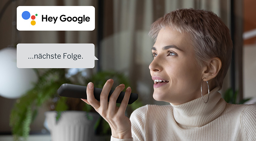 Eine Person hält ein schwarzes Gerät in der Hand, mit Textfeldern für "Hey Google" und "...nächste Folge".