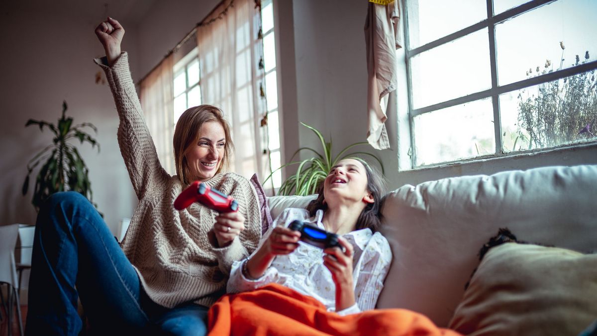 Eine Person jubelt mit rotem Controller, die andere lacht mit schwarzem auf einem Sofa. Eine Pflanze und ein Fenster sind im Hintergrund sichtbar.