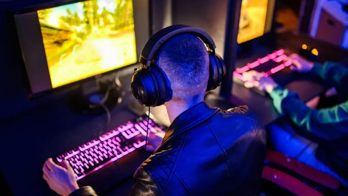 Zwei Spieler im dunklen Raum: Einer mit Kopfhörern und schwarzer Jacke nutzt rosafarbene Tastatur vor hellem Gaming-Monitor. Rechts weiterer Spieler.