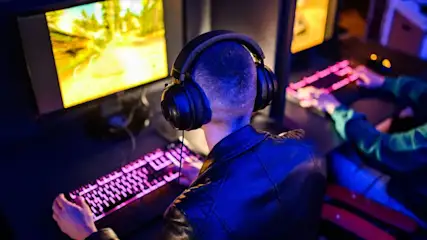 Zwei Spieler im dunklen Raum: Einer mit Kopfhörern und schwarzer Jacke nutzt rosafarbene Tastatur vor hellem Gaming-Monitor. Rechts weiterer Spieler.