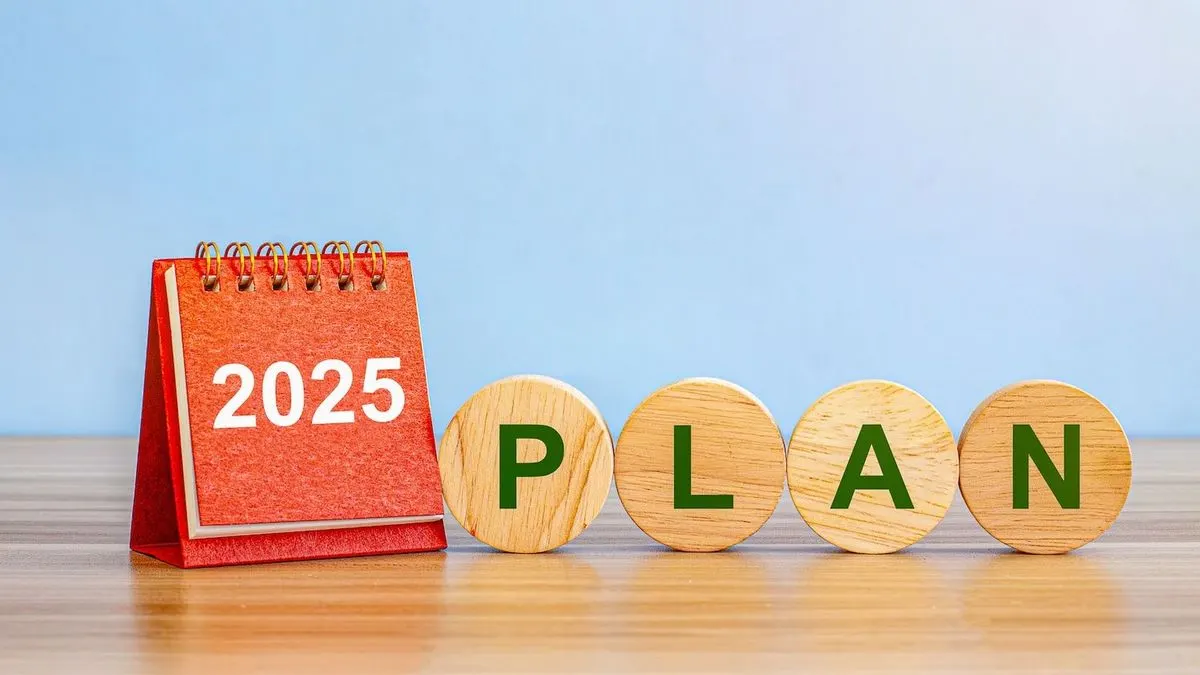 Roter Tischkalender mit "2025" und vier Holzscheiben mit grünen Buchstaben "PLAN" auf Holztisch vor blauem Hintergrund.