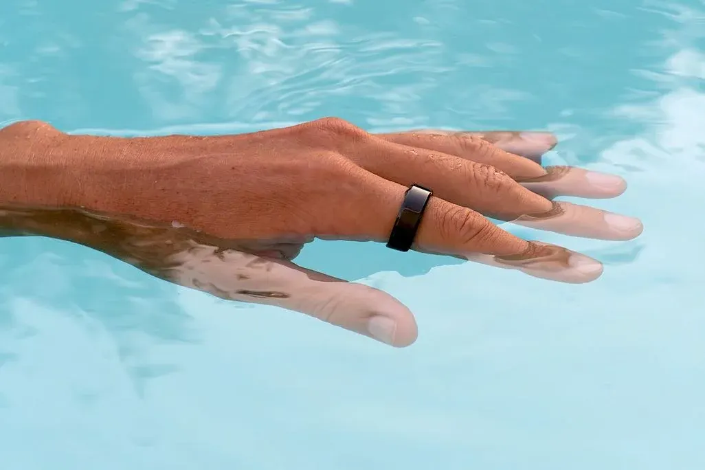 Nahaufnahme einer Hand, teilweise im Poolwasser, trägt einen schwarzen Smart‑Ring am Ringfinger.