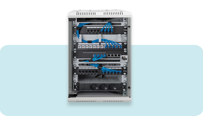 Frontansicht eines kompakten, geöffneten Wandgehäuses (Netzwerkschrank). Im Inneren sind IT-Komponenten sauber verbaut: Patchpanel und ein Netzwerk-Switch sind durch blaue Patchkabel ordentlich miteinander verbunden. Ganz unten im Schrank ist eine schwarze Steckdosenleiste mit drei Schuko-Steckdosen montiert.